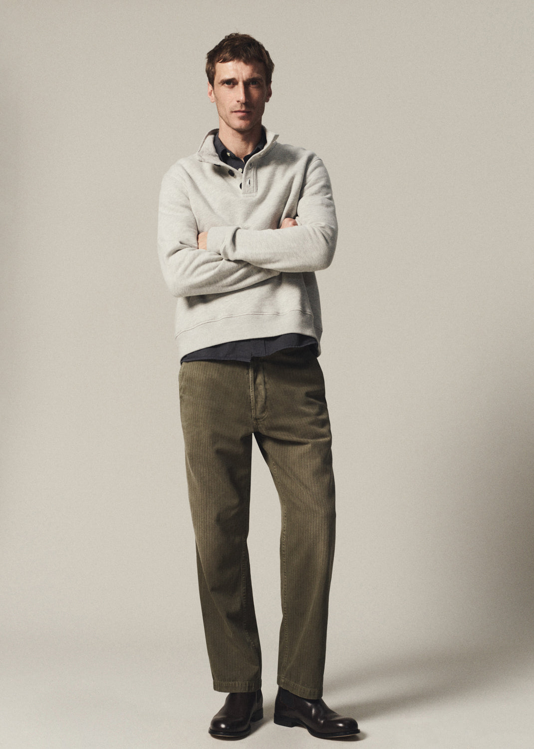 Man wearing a heather grey half-button pullover layered over a navy button-up shirt with olive pants on a grey background.
