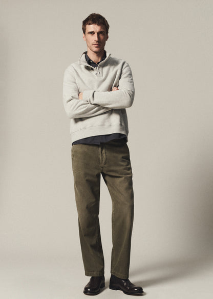 Man wearing a heather grey half-button pullover layered over a navy button-up shirt with olive pants on a grey background.