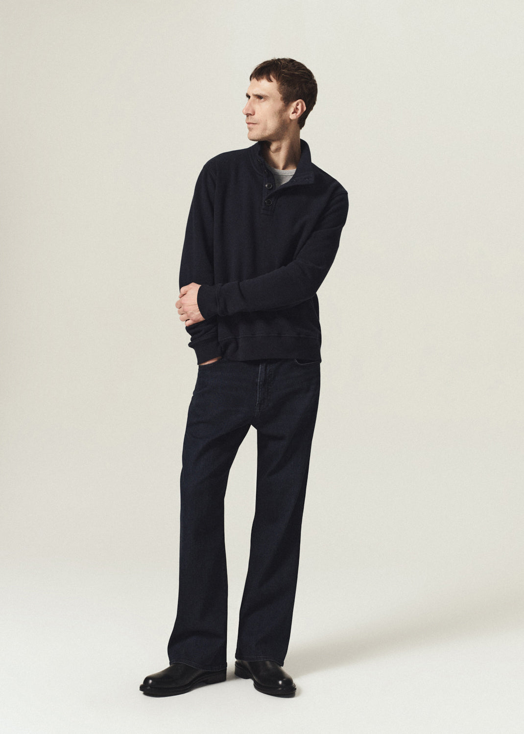 Man wearing a navy half-button pullover with dark indigo jeans and black boots on a grey background