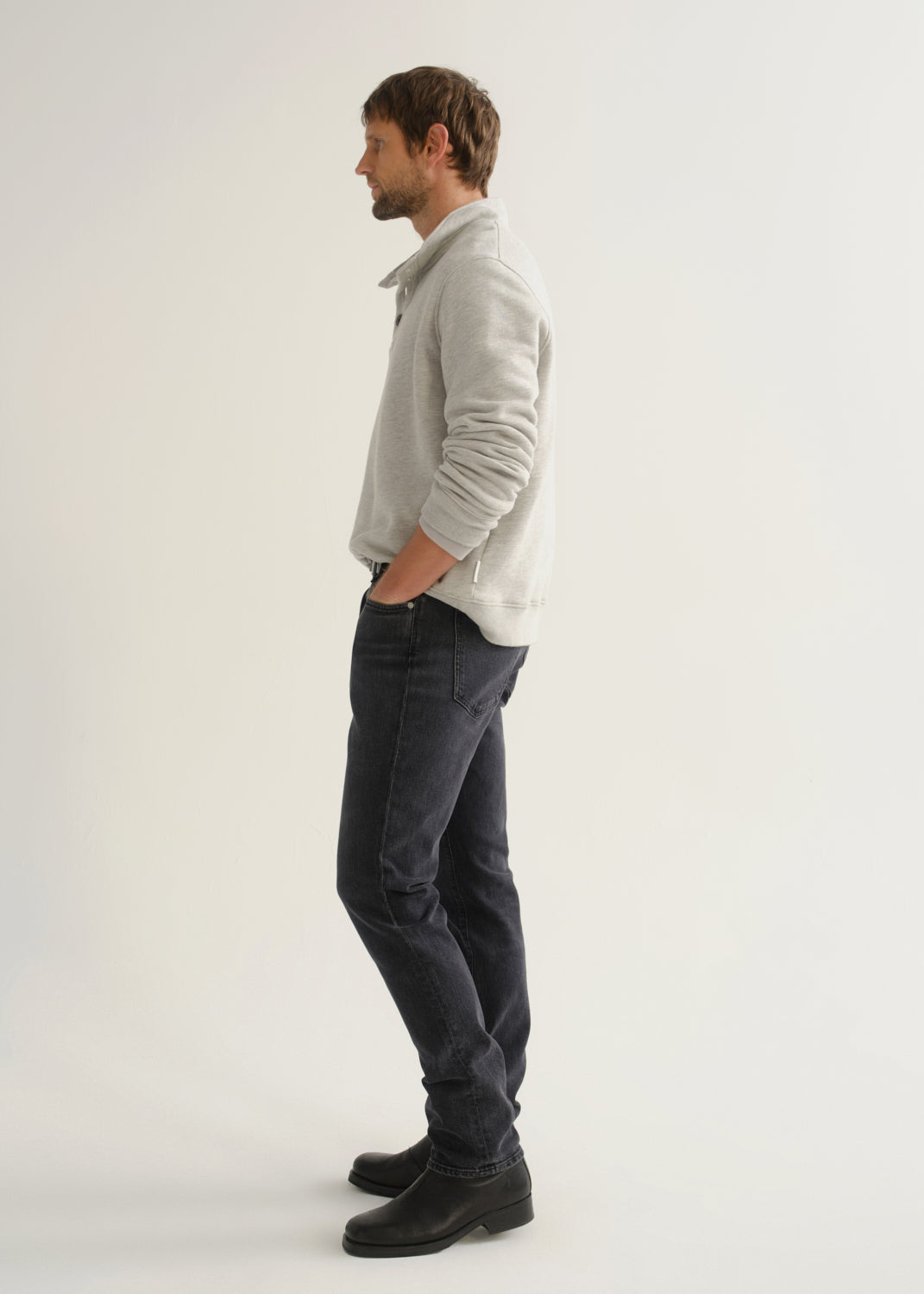Person wearing a light grey long sleeve polo pullover, black jeans, and black boots, side full body view on a light grey background.
