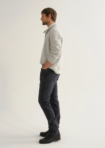 Person wearing a light grey long sleeve polo pullover, black jeans, and black boots, side full body view on a light grey background.