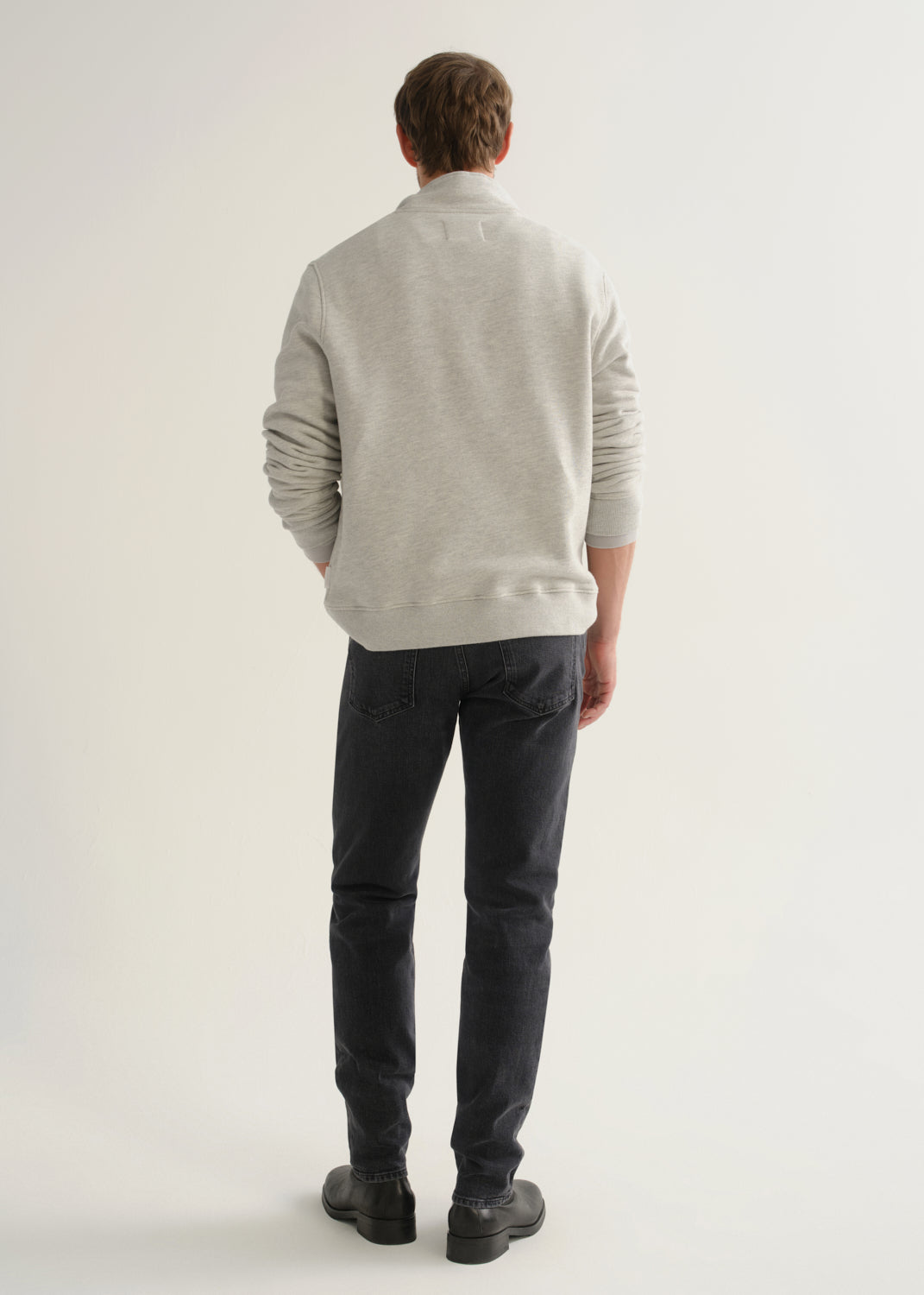 Person wearing a light grey long sleeve polo pullover, black jeans, and black boots, back full body view on a light grey background.