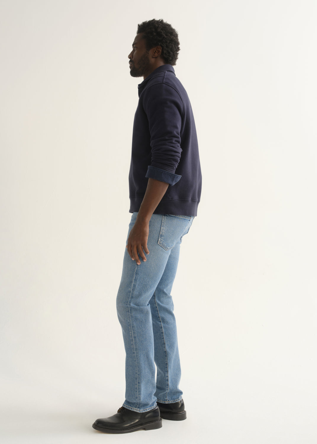 Person wearing a navy blue long sleeve pullover with light blue jeans and black shoes, side full body view on a light grey background.