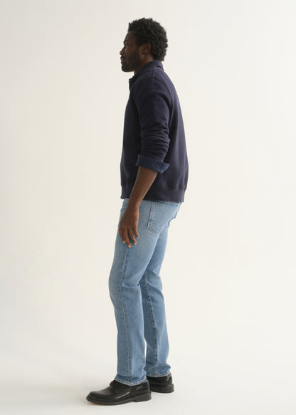 Person wearing a navy blue long sleeve pullover with light blue jeans and black shoes, side full body view on a light grey background.