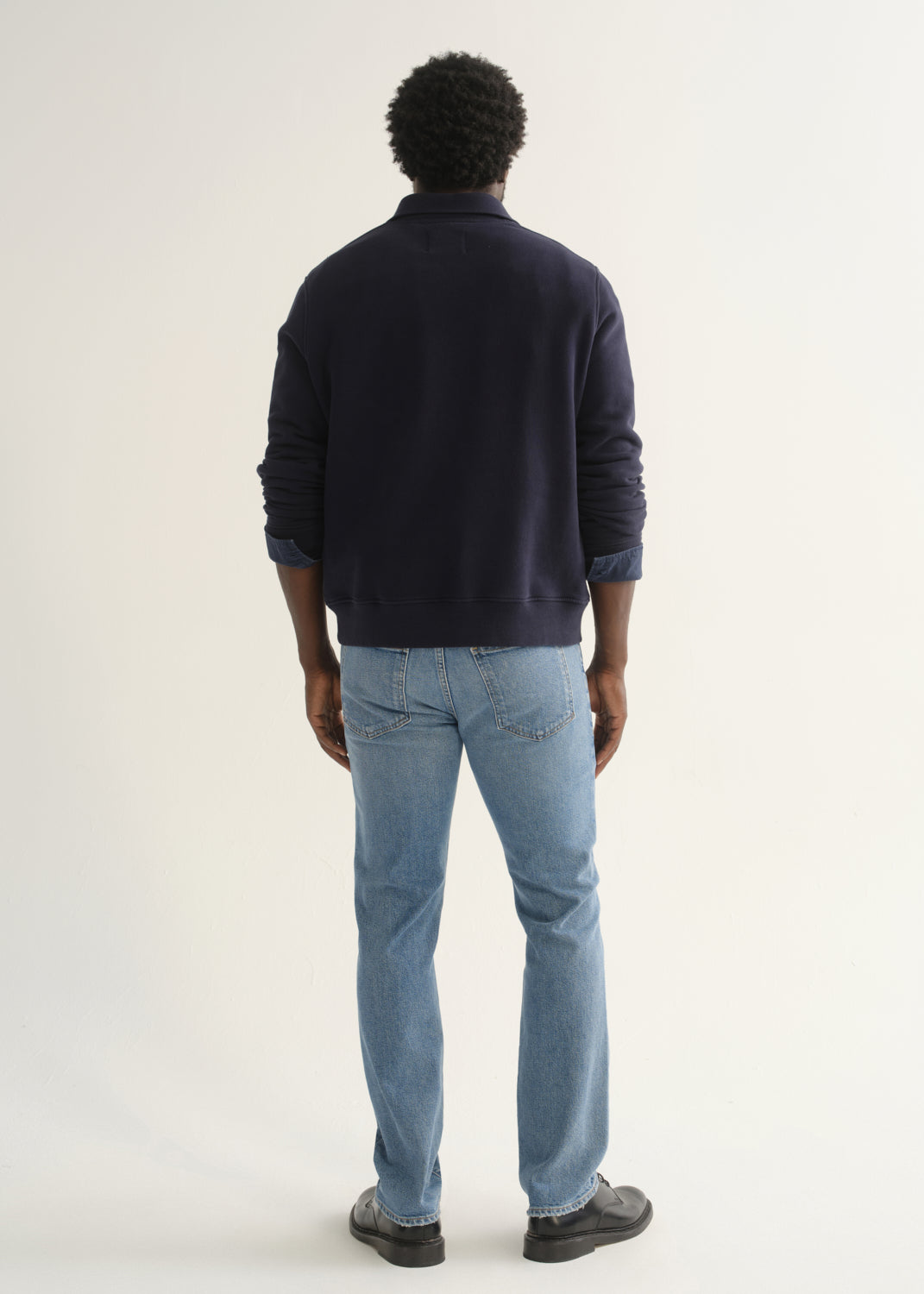 Person wearing a navy blue long sleeve pullover with light blue jeans and black shoes, back full body view on a light grey background.
