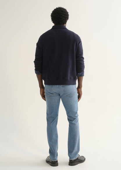 Person wearing a navy blue long sleeve pullover with light blue jeans and black shoes, back full body view on a light grey background.