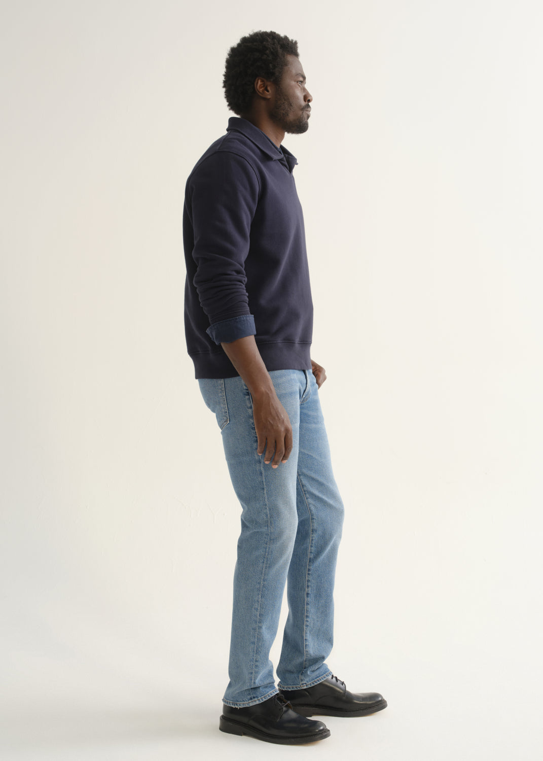 Person wearing a navy blue long sleeve pullover with light blue jeans and black shoes, angled front full body view on a light grey background.
