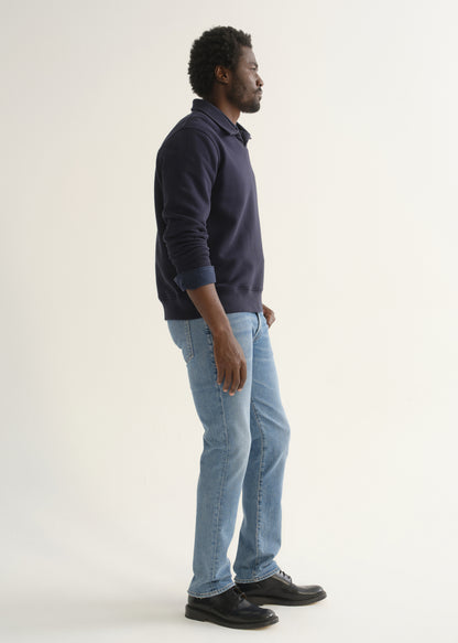Person wearing a navy blue long sleeve pullover with light blue jeans and black shoes, angled front full body view on a light grey background.