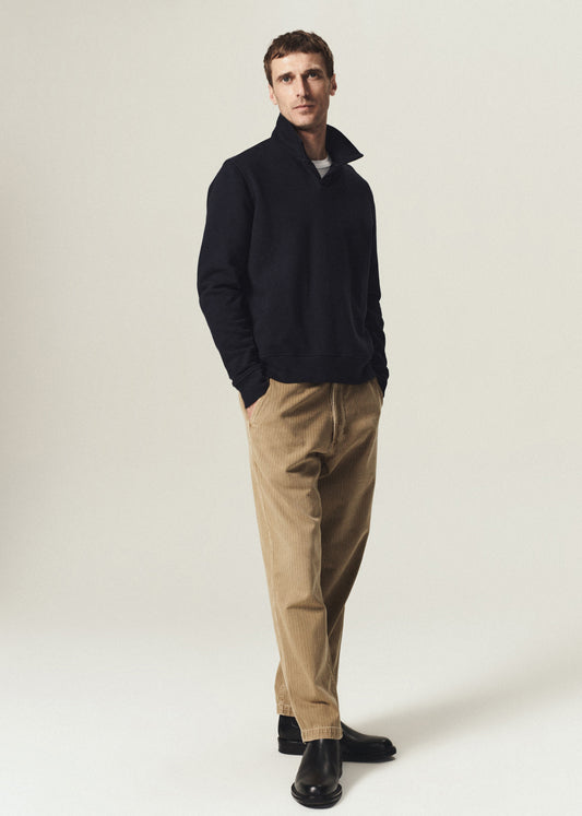 Man wearing a navy rugby pullover with tan corduroy pants and black boots on a grey background