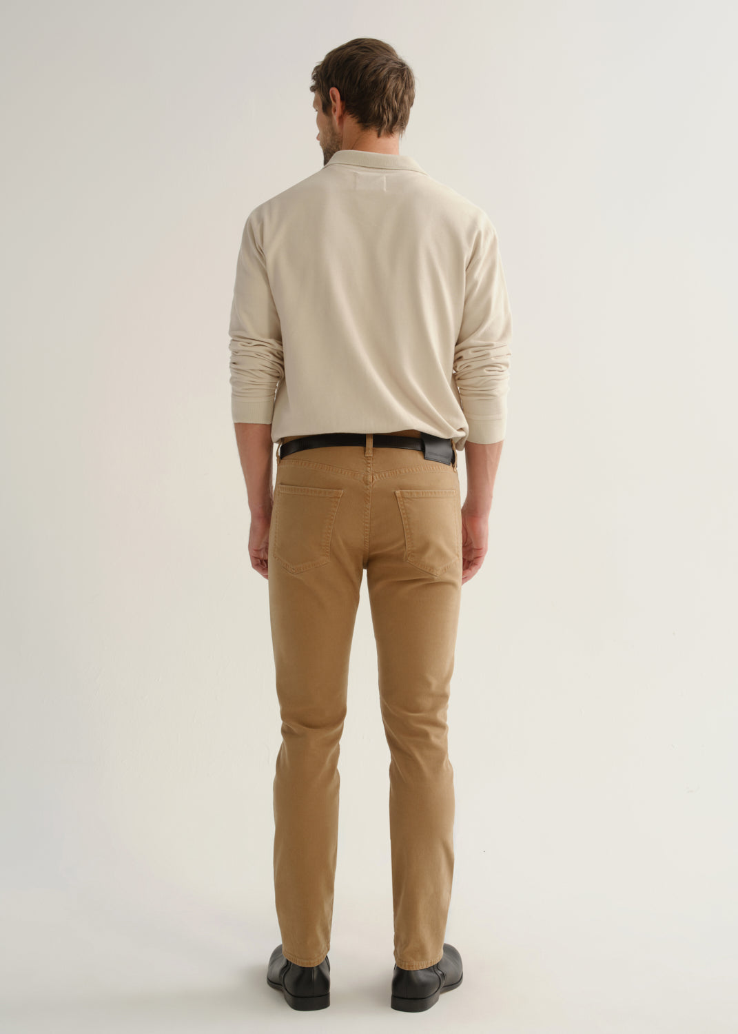 Person wearing a cream long sleeve polo pullover, tan pants, and black boots, back full body view on a light grey background.