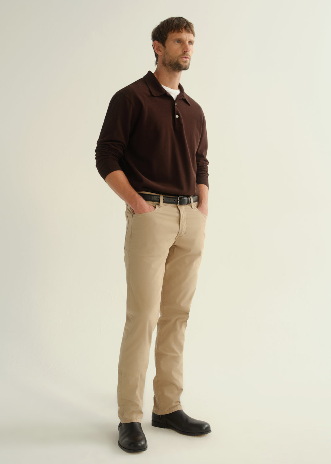 Person wearing a dark brown long sleeve polo pullover, beige pants, and black boots, angled front full body view on a light grey background.