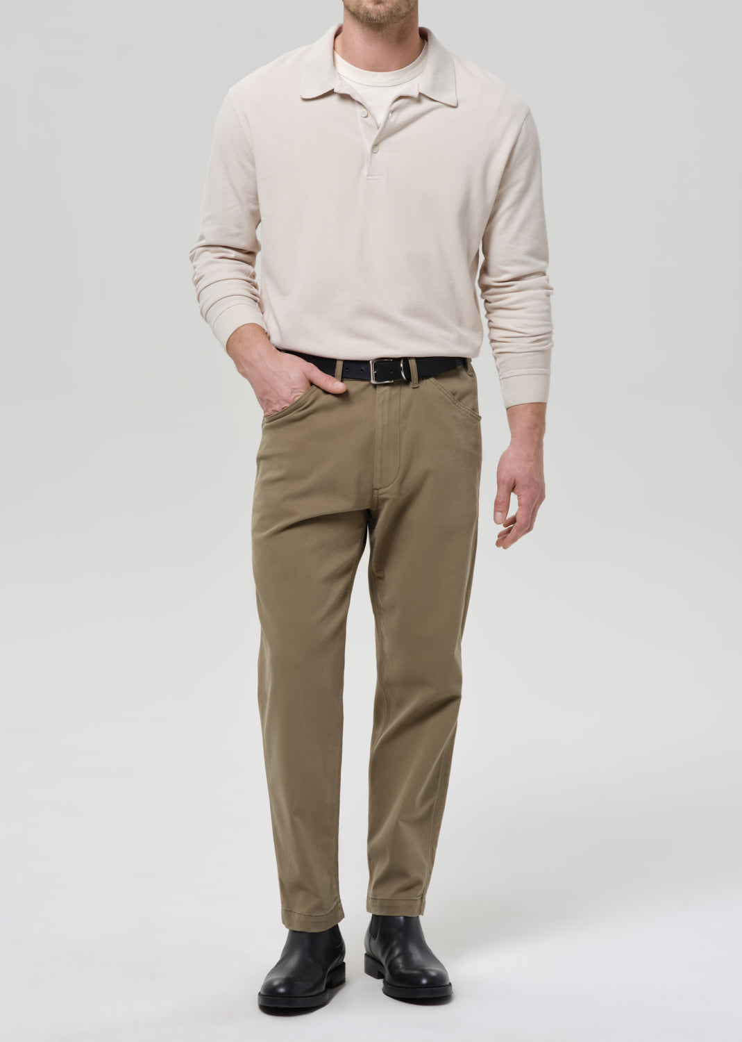 Man wearing a beige polo sweater, brown pants, and black shoes on a grey background