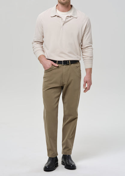 Man wearing a beige polo sweater, brown pants, and black shoes on a grey background