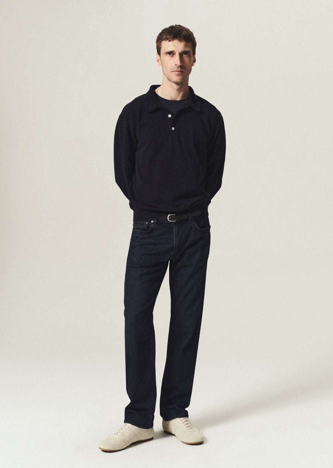 Man wearing a navy long sleeve polo with dark indigo jeans and cream sneakers on a grey background